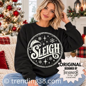 Könnte beinhalten: Ein schwarzes Langarm-Sweatshirt mit einer großen, weißen, kreisförmigen Grafik in Used-Optik. Das Design zeigt das Wort SLEIGH in einer stilisierten Schriftart, eine Weihnachtsmannmütze, Schneeflocken und Sterne. Ein weißes sternförmiges Etikett zeigt Text.