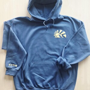 K&ouml;nnte beinhalten: Ein schieferblauer Kapuzenpullover mit einer gelben Grafik einer aufgehenden Sonne &uuml;ber Bergen auf der linken Brust. Der Text "Moses 6:34" ist in Gelb auf dem rechten &Auml;rmelb&uuml;ndchen aufgedruckt. Das Kleidungsst&uuml;ck hat eine K&auml;ngurutasche vorne.