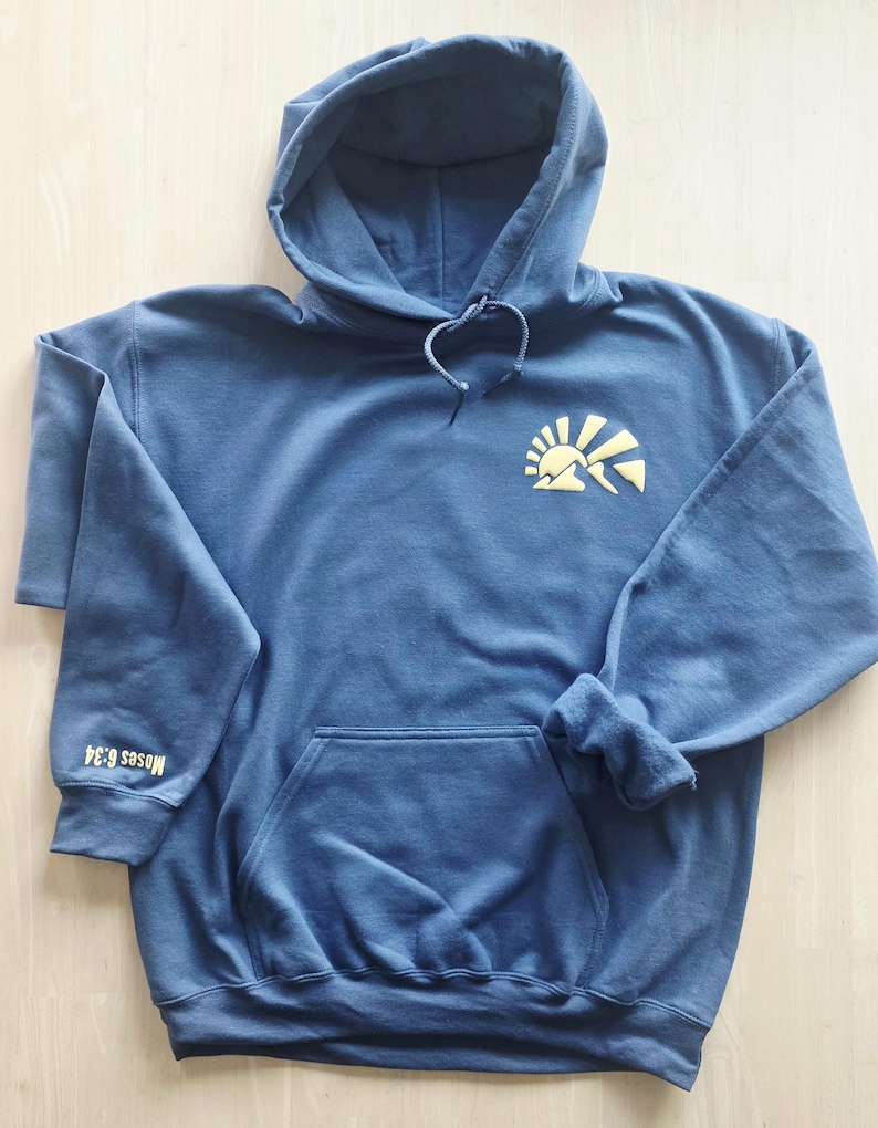 K&ouml;nnte beinhalten: Ein schieferblauer Kapuzenpullover mit einer gelben Grafik einer aufgehenden Sonne &uuml;ber Bergen auf der linken Brust. Der Text "Moses 6:34" ist in Gelb auf dem rechten &Auml;rmelb&uuml;ndchen aufgedruckt. Das Kleidungsst&uuml;ck hat eine K&auml;ngurutasche vorne.