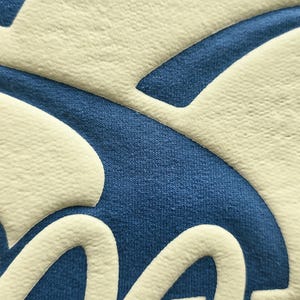 K&ouml;nnte beinhalten: Eine Nahaufnahme eines blauen Kleidungsst&uuml;cks mit einem erhabenen, cremefarbenen Grafikdesign. Die Textur des Textils ist sichtbar und zeigt eine weiche, gestrickte Oberfl&auml;che mit einem dicken, glatten und leicht erh&ouml;hten Druck.