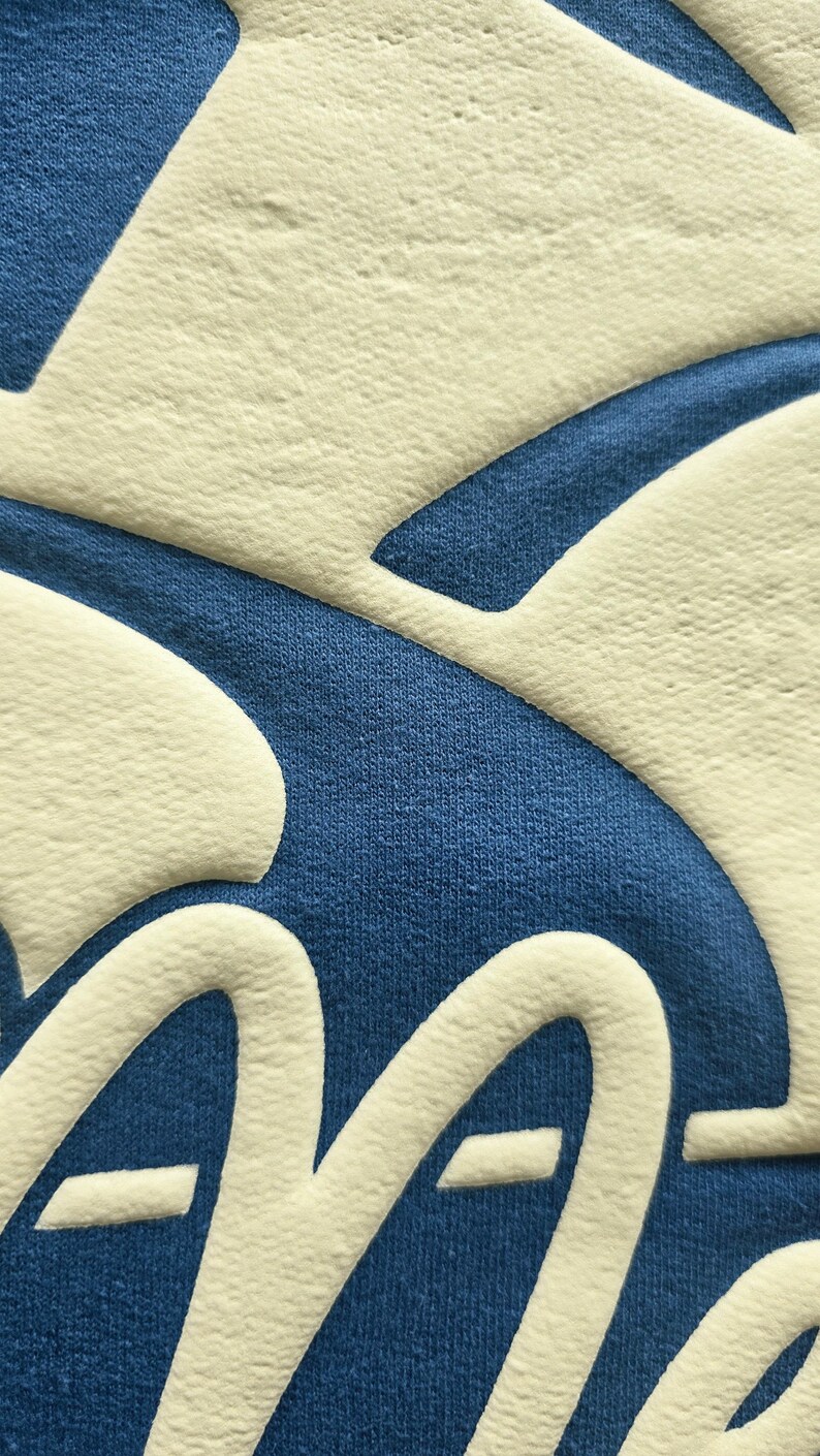 K&ouml;nnte beinhalten: Eine Nahaufnahme eines blauen Kleidungsst&uuml;cks mit einem erhabenen, cremefarbenen Grafikdesign. Die Textur des Textils ist sichtbar und zeigt eine weiche, gestrickte Oberfl&auml;che mit einem dicken, glatten und leicht erh&ouml;hten Druck.
