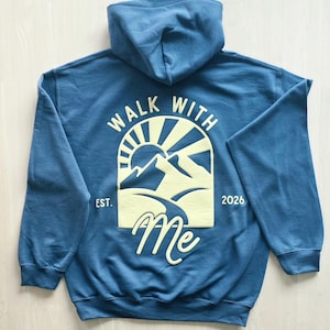 K&ouml;nnte beinhalten: Ein blauer Kapuzenpullover mit einer hellgelben Grafik auf der R&uuml;ckseite. Das Design zeigt eine Sonne &uuml;ber Bergen mit einem Pfad, den Text "WALK WITH ME" und "EST. 2026" an den Seiten. Der Kapuzenpullover hat lange &Auml;rmel und eine Kapuze.