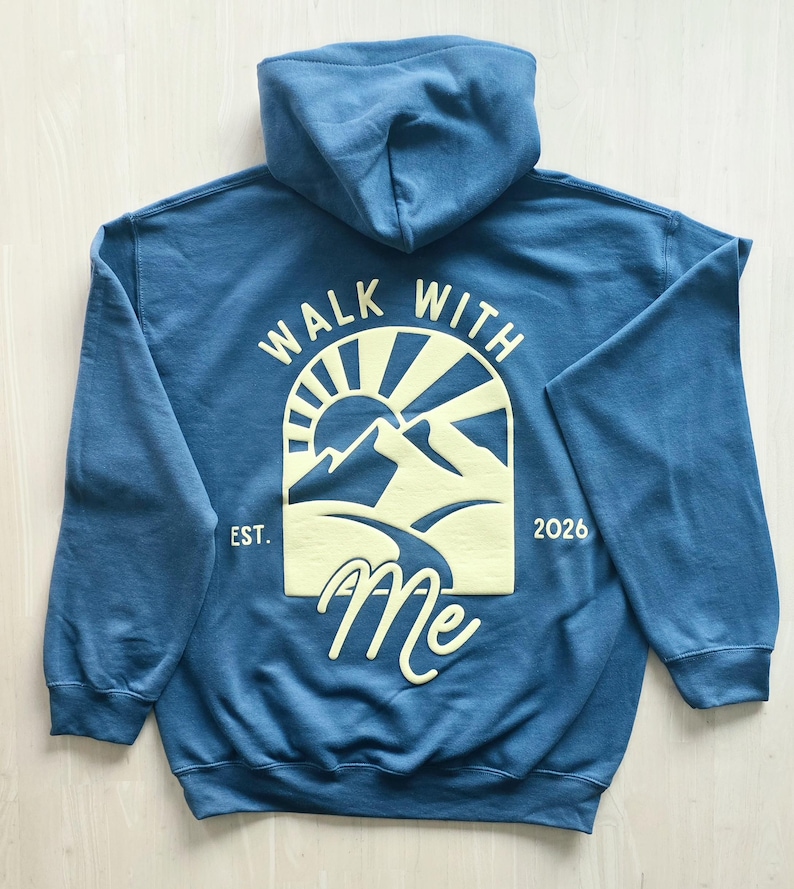 K&ouml;nnte beinhalten: Ein blauer Kapuzenpullover mit einer hellgelben Grafik auf der R&uuml;ckseite. Das Design zeigt eine Sonne &uuml;ber Bergen mit einem Pfad, den Text "WALK WITH ME" und "EST. 2026" an den Seiten. Der Kapuzenpullover hat lange &Auml;rmel und eine Kapuze.