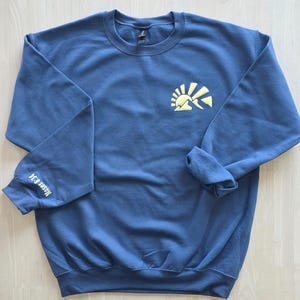 K&ouml;nnte beinhalten: Ein schieferblaues Sweatshirt mit Rundhalsausschnitt und einer hellgelben Grafik einer aufgehenden Sonne &uuml;ber Bergen auf der linken Brust. Der Text "Moses 6:34" ist in Hellgelb auf der Au&szlig;enseite des rechten &Auml;rmels aufgedruckt.