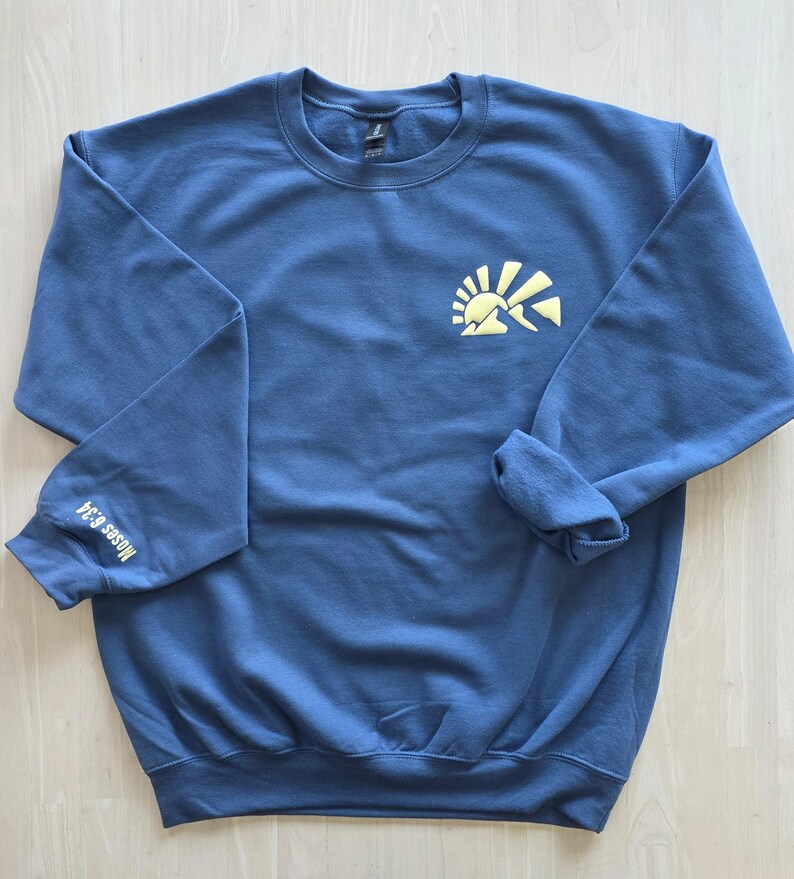 K&ouml;nnte beinhalten: Ein schieferblaues Sweatshirt mit Rundhalsausschnitt und einer hellgelben Grafik einer aufgehenden Sonne &uuml;ber Bergen auf der linken Brust. Der Text "Moses 6:34" ist in Hellgelb auf der Au&szlig;enseite des rechten &Auml;rmels aufgedruckt.