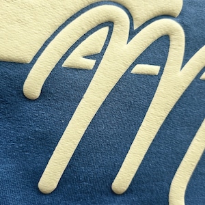 K&ouml;nnte beinhalten: Ein blaues Kleidungsst&uuml;ck aus Stoff mit einem gro&szlig;en, erhabenen, cremefarbenen Grafikdruck. Das Design zeigt einen Teil einer geschwungenen Form am oberen Rand und die Buchstaben 'AA' in einer fetten, abgerundeten Schreibschrift.