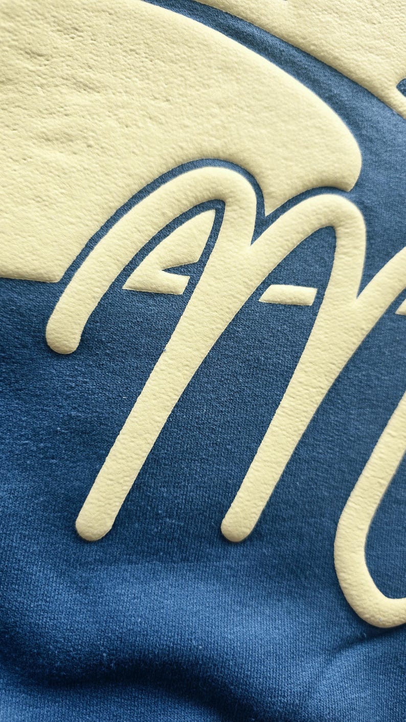 K&ouml;nnte beinhalten: Ein blaues Kleidungsst&uuml;ck aus Stoff mit einem gro&szlig;en, erhabenen, cremefarbenen Grafikdruck. Das Design zeigt einen Teil einer geschwungenen Form am oberen Rand und die Buchstaben 'AA' in einer fetten, abgerundeten Schreibschrift.