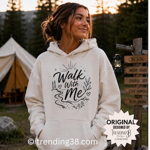 May include: A cream-colored hooded sweatshirt featuring a black graphic print on the chest. The design includes the text "Walk With Me" surrounded by illustrations of a sun, a winding path, small hearts, leaves, and flowers in a circular layout.