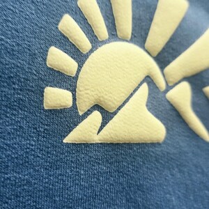 K&ouml;nnte beinhalten: Ein dunkelblaues Kleidungsst&uuml;ck aus Stoff mit einem erhabenen, strukturierten Grafikdruck in Hellgelb. Das Design zeigt eine stilisierte Sonne, die &uuml;ber einem Berggipfel aufgeht, mit ausgepr&auml;gten Strahlen, die von der Sonne ausgehen.