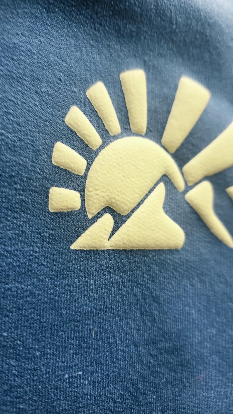 K&ouml;nnte beinhalten: Ein dunkelblaues Kleidungsst&uuml;ck aus Stoff mit einem erhabenen, strukturierten Grafikdruck in Hellgelb. Das Design zeigt eine stilisierte Sonne, die &uuml;ber einem Berggipfel aufgeht, mit ausgepr&auml;gten Strahlen, die von der Sonne ausgehen.