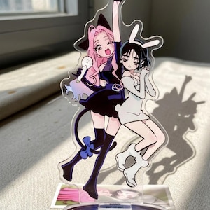 Può includere: Un supporto in acrilico trasparente con due personaggi in stile anime. Un personaggio ha i capelli rosa, un abito nero e un microfono. L'altro personaggio ha i capelli neri e un abito bianco con orecchie da coniglio. Il supporto è su una base rettangolare.