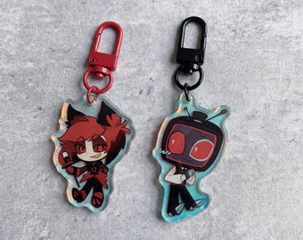 May include: Two acrylic keychains with cartoon character designs. One keychain features a red and black character with a red clasp. The other keychain has a black clasp and a character with a TV head and red eyes. Both keychains are on a light blue background.
