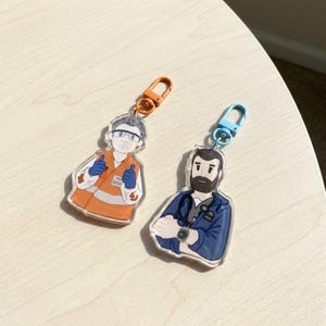 Op de afbeelding: Twee acryl sleutelhangers met geïllustreerde figuren. De ene heeft een oranje sluiting en toont een persoon in een oranje veiligheidsvest met blauwe handschoenen die een duim omhoog steekt. De andere heeft een blauwe sluiting en toont een persoon met een baard en stethoscoop.