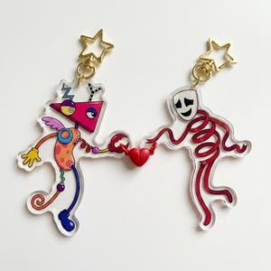 Puede incluir: Dos llaveros de acrílico con cierres dorados en forma de estrella. Uno muestra una figura robótica abstracta y colorida con cabeza triangular y una pinza. El otro es una figura roja en espiral con una máscara blanca. Sostienen un corazón rojo.