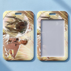 Può includere: Due porta badge rettangolari. Uno presenta un'illustrazione dettagliata di una persona in tuta bianca e una figura meccanica, su sfondo dorato. L'altro supporto è vuoto con una finestra di plastica trasparente.