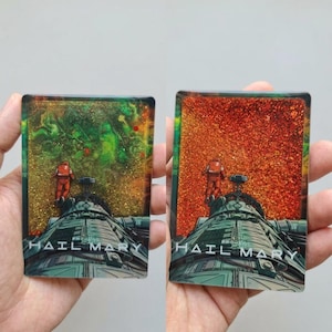 Puede incluir: Dos piezas de arte rectangulares con temática espacial. Una tiene un fondo verde y dorado, la otra un fondo naranja. Ambas muestran un astronauta y el texto "HAIL MARY" en una nave espacial.