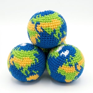 May include: A stack of three crocheted balls, each depicting a globe. The balls feature blue oceans, green and yellow continents, and white accents. The handmade spheres are arranged against a white background.
