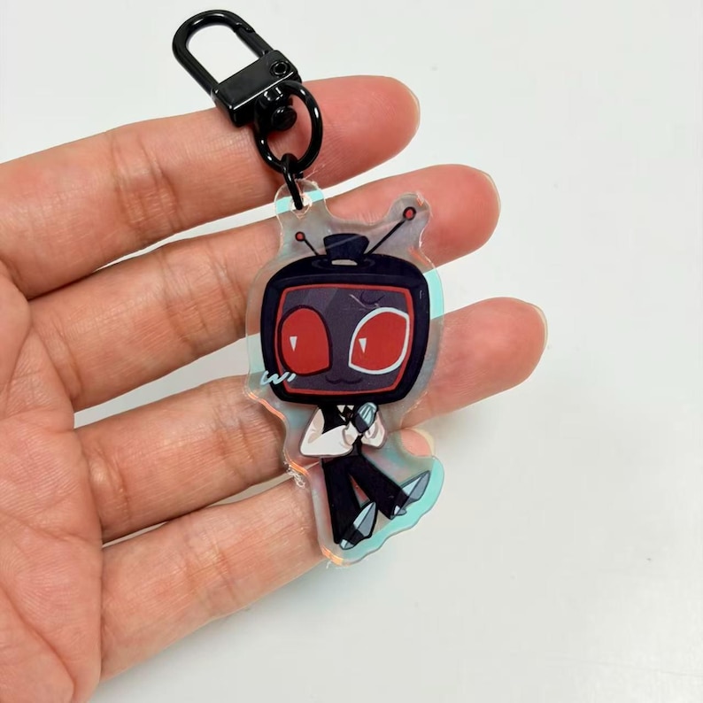 May include: A clear acrylic keyring featuring a cartoon character with a black television head, red screen, and two antennas. The character wears a white shirt and black trousers, attached to a black metal lobster clasp hardware.