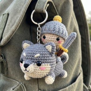 May include: Crocheted keychain featuring a gray wolf with a knight riding on its back. The knight wears a gray hat and holds a sword. The wolf has a black nose and pink cheeks. The keychain is attached to a silver ring.