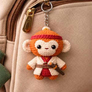 Pode incluir: Um chaveiro de macaco de crochê nas cores laranja, vermelho e creme. O macaco usa uma faixa vermelha, uma jaqueta branca e um cinto marrom com uma pequena espada. O chaveiro é preso a uma corrente prateada e um zíper dourado.