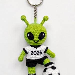 Op de afbeelding: Een gehaakte alien sleutelhanger met een groen buitenaards figuur in een wit shirt met de zwarte tekst 2026 en een zwarte broek. Het figuur heeft grote zwarte ogen en staat met één voet op een zwart-witte voetbal.