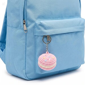 May include: A light blue backpack with a front pocket and a silver zipper. A pink crocheted macaron keychain with white and yellow sprinkles hangs from the zipper. The keychain is round and has a white filling.