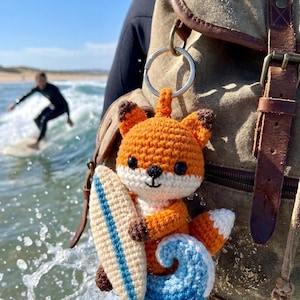 May include: A crocheted fox keyring, featuring an orange body, white face, and brown details, clutching a surfboard and a blue wave. The fox is attached to a silver keyring. A surfer rides a wave in the background.