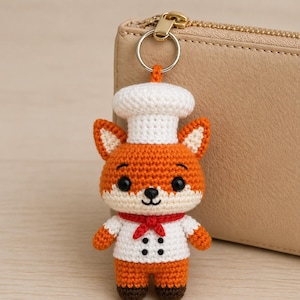 May include: A crocheted fox keychain wearing a chef's hat and uniform. The fox is orange, white, and red, with black eyes and a small black nose. The keychain has a silver ring attached to the top. The background includes a tan wallet.