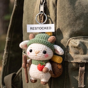 May include: A crocheted sheep keychain with a green hat, scarf, and backpack, holding a wooden stick and a red apple. The keychain is attached to a green backpack with a zipper. A white tag reads "RESTOCKED".