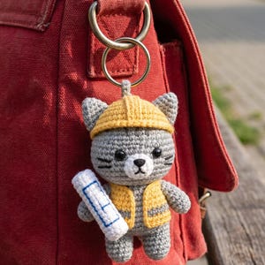 May include: A handmade crochet cat keyring in grey, yellow, and white, wearing a construction hat and vest, holding a blueprint. The cat is attached to a red canvas bag with a metal ring.