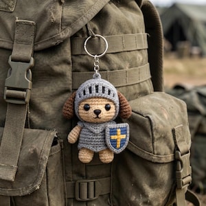 May include: A crocheted dog keychain wearing a knight's helmet and armor, holding a shield with a yellow cross on a blue background. The dog has brown ears and a tan face. The keychain is attached to a silver ring and hangs against a green backpack.