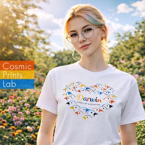 以下が含まれることがあります： カラフルな鳥のハート型デザインの白いTシャツ。「Darwin」の文字は金色の筆記体で、下に「Force of Evolution」と書かれています。庭にいる人が着ています。
