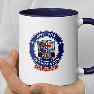 Peut inclure: Mug en céramique blanche avec intérieur et anse bleu marine. Le mug présente un motif circulaire avec le texte "ANTI-VAX ANTI-PEOPLE CLUB" et un graphique d'un bouclier brisé avec une croix médicale. Les mots "FEAR WINS SCIENCE" et "FACTS NOT INCLUDED" sont également présents.