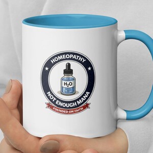 May include: White ceramic mug with a blue interior and handle. The mug features a circular graphic with the words "Homeopathy" and "Not Enough Mana" surrounding an illustration of a dropper bottle with "H2O" inside.