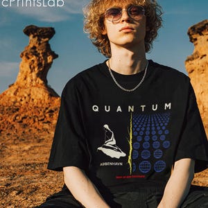 Può includere: T-shirt nero con la scritta "QUANTUM" a blocchi bianchi. La maglietta presenta un design grafico con una figura seduta e la scritta "KØBENHAVN" in bianco. La maglietta è abbinata a pantaloni cargo neri.