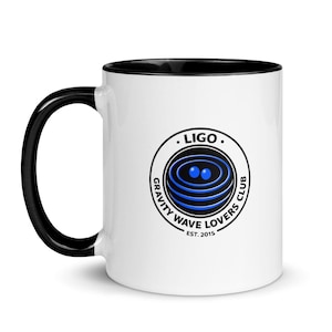 Puede incluir: Taza de cerámica blanca con asa e interior negros. La taza presenta un logotipo circular con el texto ". LIGO . GRAVITY WAVE LOVERS CLUB EST. 2015" alrededor de un gráfico azul y negro de círculos concéntricos y dos esferas azules.