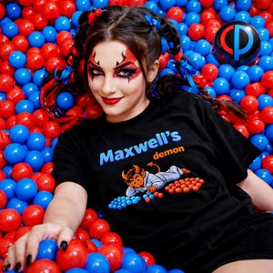 May include: Black t-shirt with the text "Maxwell's demon" in blue and orange, featuring a cartoon devil illustration. The background is a sea of red and blue plastic balls.