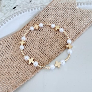 May include: Gold beaded bracelet with white faux pearls and gold cross charms. The bracelet is presented on a burlap surface. This delicate piece of jewellery is a stylish accessory. The bracelet is a delicate piece of jewellery.