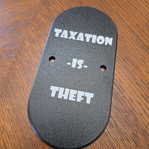 May include: A black, oval-shaped item with the white text "TAXATION -IS- THEFT". It features two small holes and is placed on a wooden surface. The text is in a bold, sans-serif font.