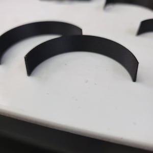 Puede incluir: Primer plano de varias piezas negras semicirculares dispuestas sobre una superficie blanca. Las piezas parecen estar hechas de un material mate y están espaciadas uniformemente. La iluminación es brillante, resaltando las formas y texturas.