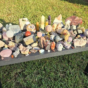 May include: A collection of various crystals and gemstones displayed on a black table. The assortment includes quartz, rose quartz, amethyst, and other colorful stones in different shapes and sizes. The table is set on a grassy surface.