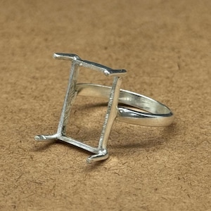 Op de afbeelding: Een zilverkleurige ring met een rechthoekige zetting, ontworpen om een edelsteen te bevatten. De zetting heeft vier pootjes en is bevestigd aan een eenvoudige band. De ring wordt getoond op een bruin oppervlak.