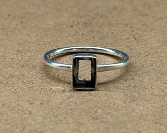 May include: A silver ring featuring a rectangular, open-centre design. The ring band is thin and circular, with the rectangular element centrally placed. The ring is set against a neutral, brown background.