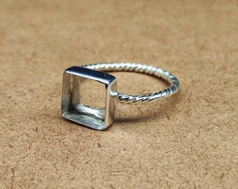 May include: A silver ring featuring a square setting and a twisted band. The ring is made of a shiny metal and is set against a brown background. The square setting is empty, ready for a gemstone or other embellishment.
