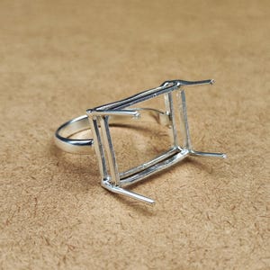 Op de afbeelding: Een zilveren ringzetting met een rechthoekig frame ontworpen om een edelsteen vast te houden. De ring heeft een gladde, gepolijste band en vier pinnen die zich uitstrekken vanaf het frame om de steen vast te zetten. De zetting staat op een neutrale achtergrond.