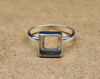 May include: A silver ring with a square setting. The ring features a polished band and a square bezel designed to hold a gemstone or other decorative element. The ring is shown on a textured, light brown surface.