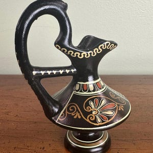 May include: A black ceramic vase with a curved handle and decorative patterns. The vase features a flared top and a detailed base. The design includes white, red, and gold accents, with a floral motif on the body.
