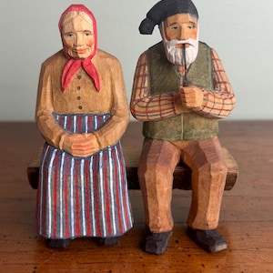 May include: Hand-carved wooden figurines of an elderly couple seated on a bench. The woman wears a red headscarf and striped skirt. The man has a beard, a hat, and holds a pipe. The figures are painted in natural wood tones.