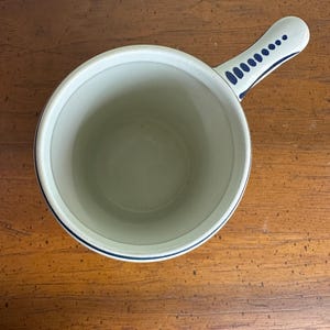 Op de afbeelding: Een witte keramische steelpan met een blauwe rand en handvat. Het handvat is versierd met een reeks blauwe stippen. De binnenkant van de steelpan is lichtbeige.