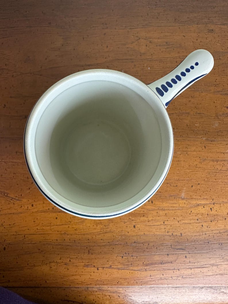 Op de afbeelding: Een witte keramische steelpan met een blauwe rand en handvat. Het handvat is versierd met een reeks blauwe stippen. De binnenkant van de steelpan is lichtbeige.